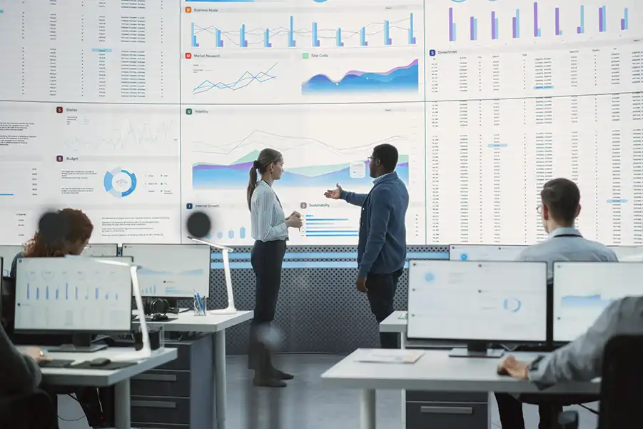 Two people in front of large screens with financial graphs, illustrating the impact on business operations.